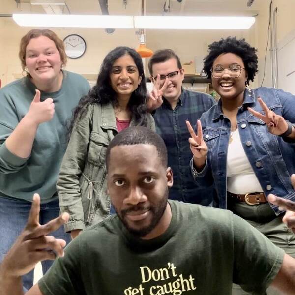 Five individuals smile playfully in a lab setting. A man in a dark green "Don't get caught with your plants down" shirt is in the foreground with a hand gesture. Behind him, a woman gives a thumbs-up and two others flash peace signs, all grinning.
