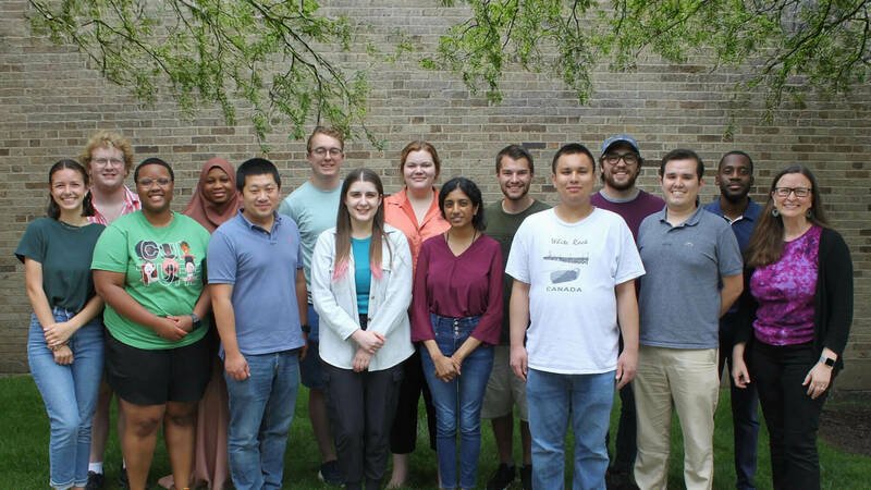 A diverse group of 14 smiling men and women of varying ages stand outdoors against a light brown brick wall.